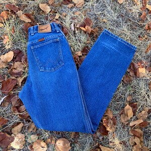 Pu&ograve; includere: Jeans Wrangler in denim blu con un classico design a cinque tasche. I jeans presentano una toppa in pelle marrone con il logo Wrangler. I jeans sono di un lavaggio medio e sono appoggiati su erba secca e foglie cadute.