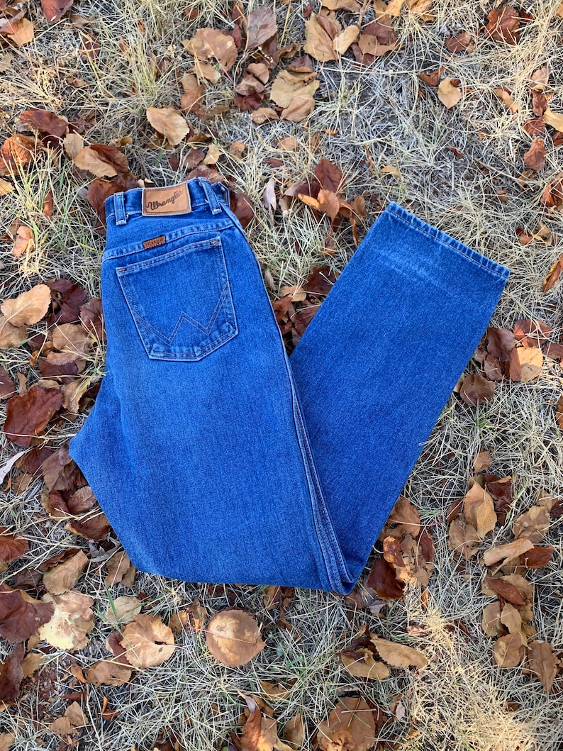 Pu&ograve; includere: Jeans Wrangler in denim blu con un classico design a cinque tasche. I jeans presentano una toppa in pelle marrone con il logo Wrangler. I jeans sono di un lavaggio medio e sono appoggiati su erba secca e foglie cadute.