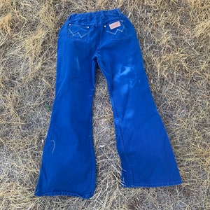 May include: Dark blue Wrangler jeans with a flared leg design. The jeans feature white stitching on the back pockets and a brown leather patch with the Wrangler logo. The jeans are laid out on a bed of dry grass.