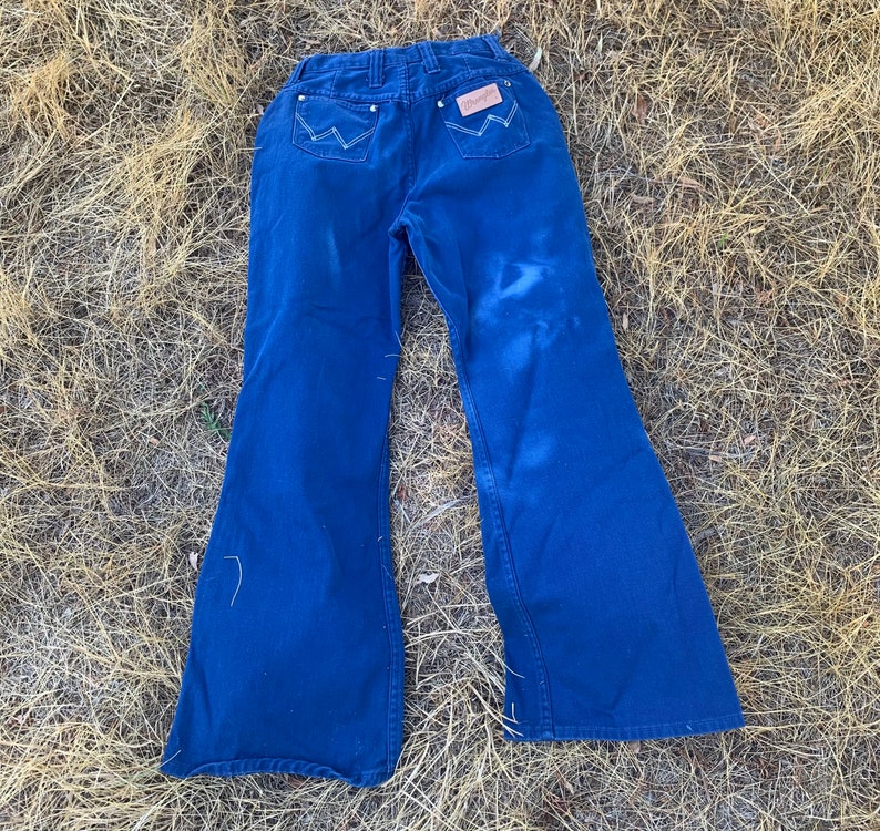 May include: Dark blue Wrangler jeans with a flared leg design. The jeans feature white stitching on the back pockets and a brown leather patch with the Wrangler logo. The jeans are laid out on a bed of dry grass.