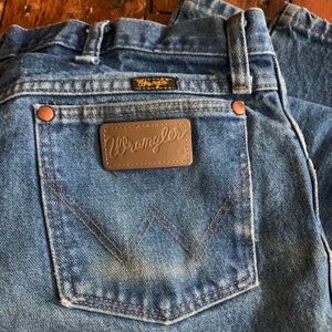 May include: Close-up of blue denim jeans with a Wrangler label on the back pocket. The jeans have copper rivets and a classic five-pocket design. The Wrangler logo is visible on the back pocket and above the pocket.