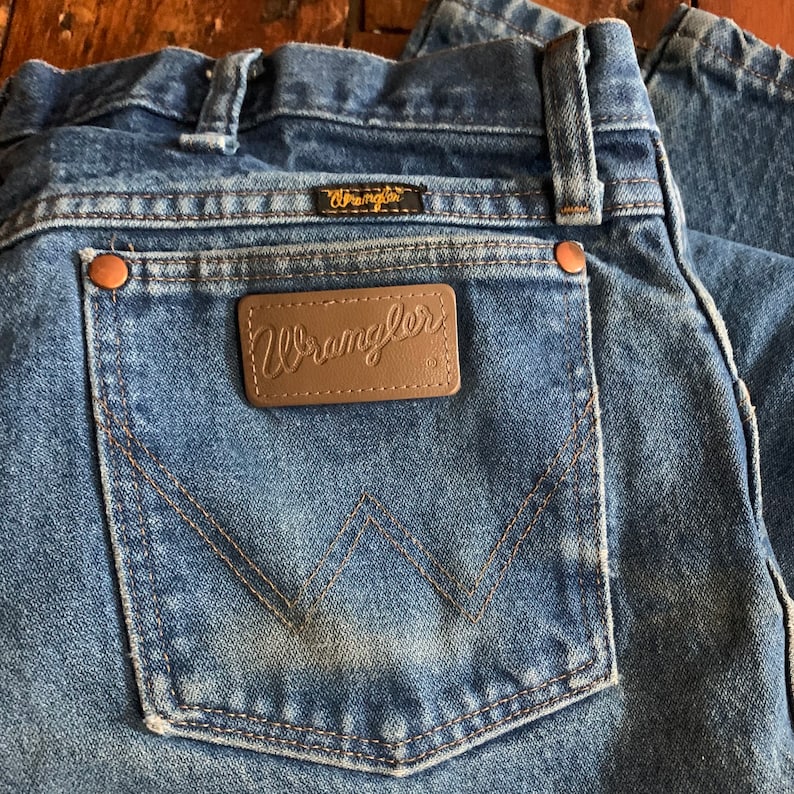 May include: Close-up of blue denim jeans with a Wrangler label on the back pocket. The jeans have copper rivets and a classic five-pocket design. The Wrangler logo is visible on the back pocket and above the pocket.