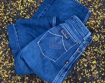 Può includere: Jeans di denim blu scuro con vita alta e gamba svasata. La tasca posteriore presenta cuciture bianche e un'etichetta con la scritta "Bon Jour". I jeans sono piegati e appoggiati su una superficie ricoperta di fiori gialli.
