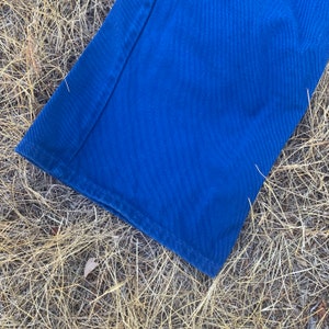 May include: Close-up of a section of blue denim pants. The fabric is a vibrant shade of blue, with visible stitching along the seams and hem. The pants are lying on a bed of dry, golden-brown grass.