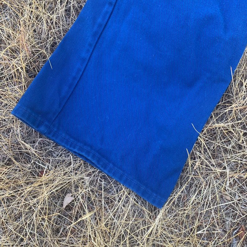 May include: Close-up of a section of blue denim pants. The fabric is a vibrant shade of blue, with visible stitching along the seams and hem. The pants are lying on a bed of dry, golden-brown grass.