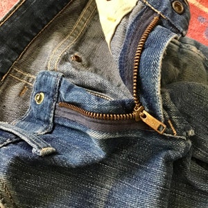 May include: Close-up of blue denim jeans with a partially open brass zipper. The jeans show signs of wear and fading, with visible stitching and metal rivets. The zipper pull has the text "0072 C".
