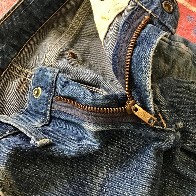 May include: Close-up of blue denim jeans with a partially open brass zipper. The jeans show signs of wear and fading, with visible stitching and metal rivets. The zipper pull has the text "0072 C".