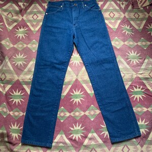 May include: Dark blue denim jeans with a classic five-pocket design. The jeans have a straight leg cut and a button and zipper closure. The denim is a solid, medium-weight fabric. The jeans are laid flat on a patterned fabric.