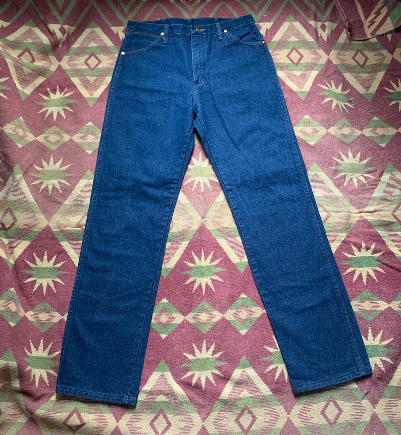 May include: Dark blue denim jeans with a classic five-pocket design. The jeans have a straight leg cut and a button and zipper closure. The denim is a solid, medium-weight fabric. The jeans are laid flat on a patterned fabric.