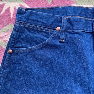 May include: Close-up of a pair of blue denim jeans. The jeans feature a classic five-pocket design with copper-colored rivets and stitching. The pocket edges are reinforced with a contrasting thread. The fabric has a slightly worn appearance.