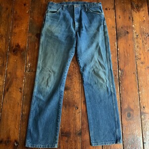 May include: A pair of blue denim jeans with a faded wash. The jeans have a classic five-pocket design, a button and zipper closure, and a straight leg. The denim has a worn appearance, with lighter areas on the thighs and knees.