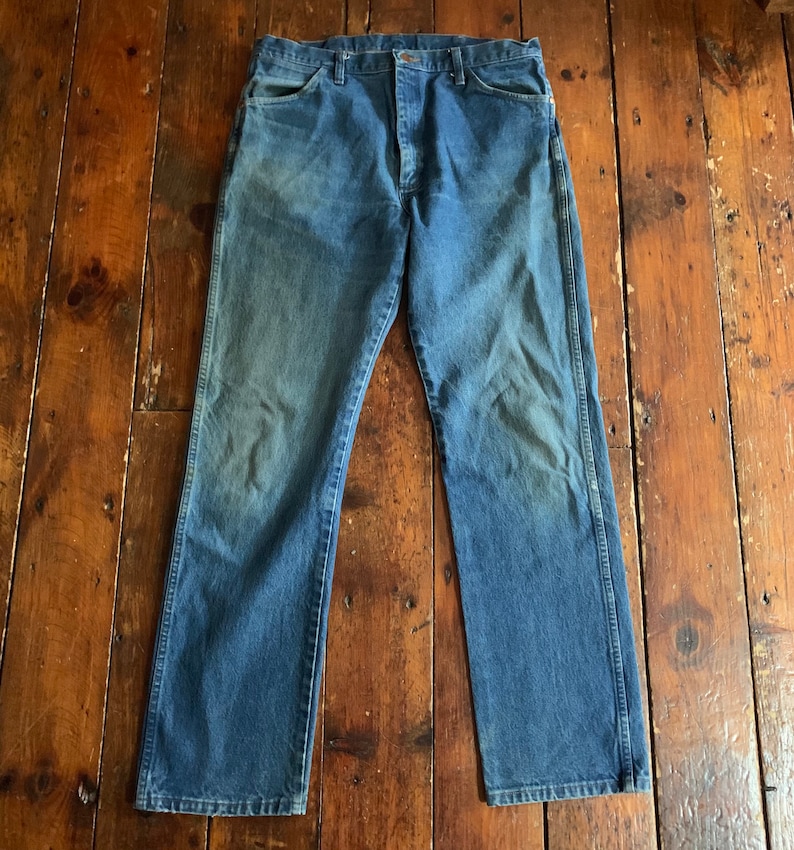 May include: A pair of blue denim jeans with a faded wash. The jeans have a classic five-pocket design, a button and zipper closure, and a straight leg. The denim has a worn appearance, with lighter areas on the thighs and knees.
