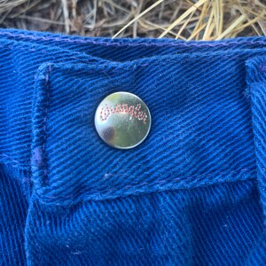 May include: Close-up of a section of blue denim jeans. A silver button with the brand name "Wrangler" in red script is visible. The jeans have a classic five-pocket design.
