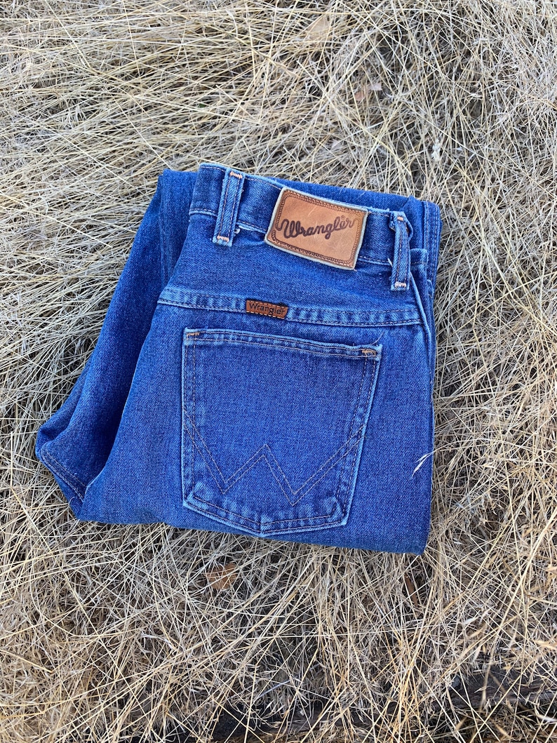 Pu&ograve; includere: Jeans blu scuro con il logo Wrangler su un'etichetta in pelle marrone. I jeans sono piegati, mostrando il design della tasca posteriore e l'etichetta Wrangler sulla tasca.