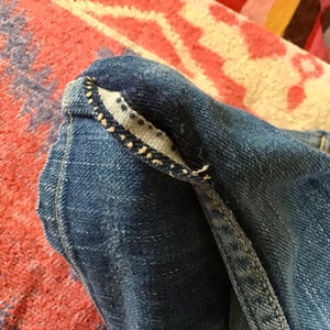 May include: Close-up of a section of blue denim jeans. The image highlights the folded hem, revealing a patterned interior lining with small, dark dots. The stitching is visible along the edges, showcasing the construction details.