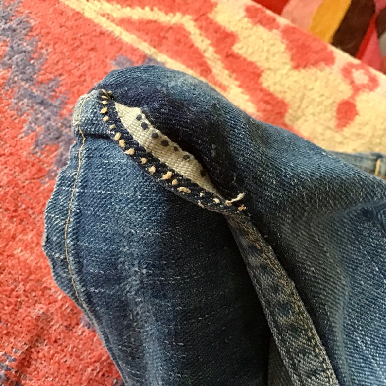 May include: Close-up of a section of blue denim jeans. The image highlights the folded hem, revealing a patterned interior lining with small, dark dots. The stitching is visible along the edges, showcasing the construction details.