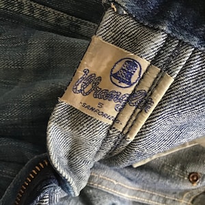 May include: Close-up of a vintage pair of blue denim jeans. The Wrangler label is visible, featuring the brand name in blue script and a bell logo. The jeans show signs of wear, with a zipper and button detail.