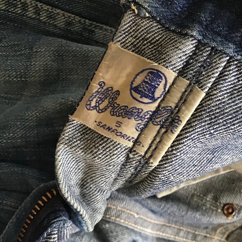 May include: Close-up of a vintage pair of blue denim jeans. The Wrangler label is visible, featuring the brand name in blue script and a bell logo. The jeans show signs of wear, with a zipper and button detail.