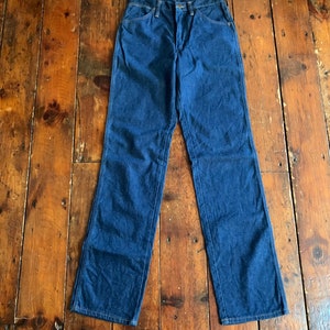 May include: A pair of dark blue denim jeans with a classic five-pocket design. The jeans have a straight leg cut and are laid flat on a wooden surface. The denim appears to be a medium weight.