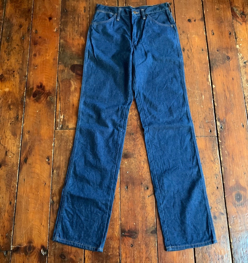 May include: A pair of dark blue denim jeans with a classic five-pocket design. The jeans have a straight leg cut and are laid flat on a wooden surface. The denim appears to be a medium weight.