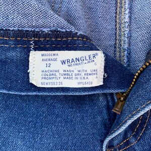 Pu&ograve; includere: Primo piano di jeans blu con un'etichetta visibile. L'etichetta recita "Wrangler" e include la taglia "12". I jeans hanno una cerniera e una cucitura visibile. L'etichetta include anche le istruzioni di lavaggio.