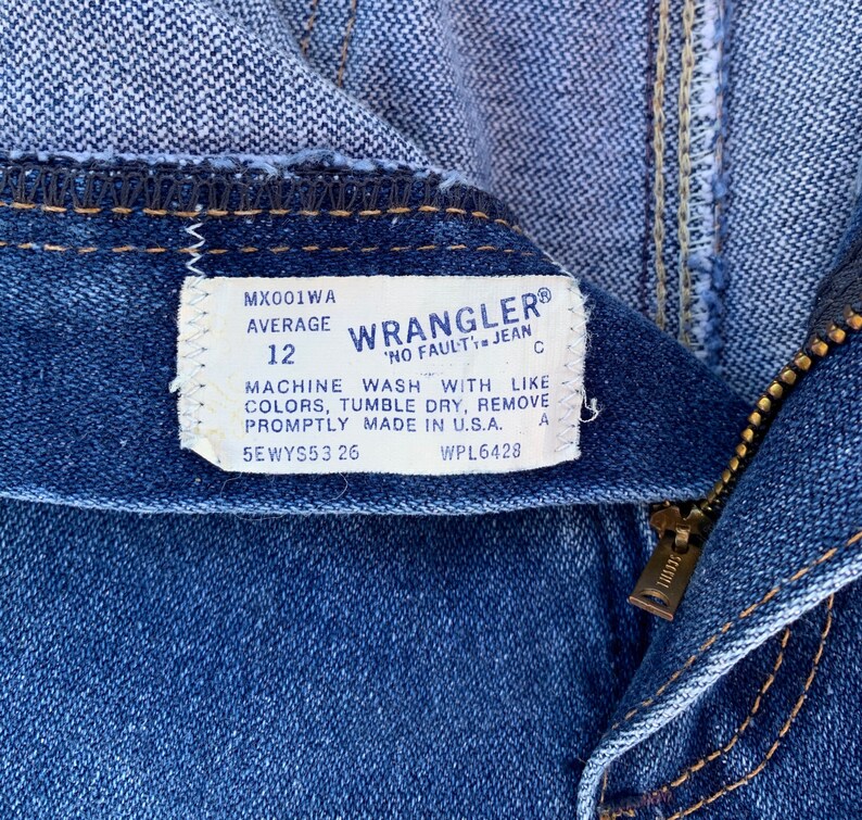 Pu&ograve; includere: Primo piano di jeans blu con un'etichetta visibile. L'etichetta recita "Wrangler" e include la taglia "12". I jeans hanno una cerniera e una cucitura visibile. L'etichetta include anche le istruzioni di lavaggio.