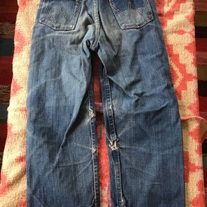 May include: Vintage blue denim jeans with the Wrangler logo on the back pockets. The jeans have a worn appearance with distressed details at the knees and hems. The jeans are laid flat on a patterned textile.