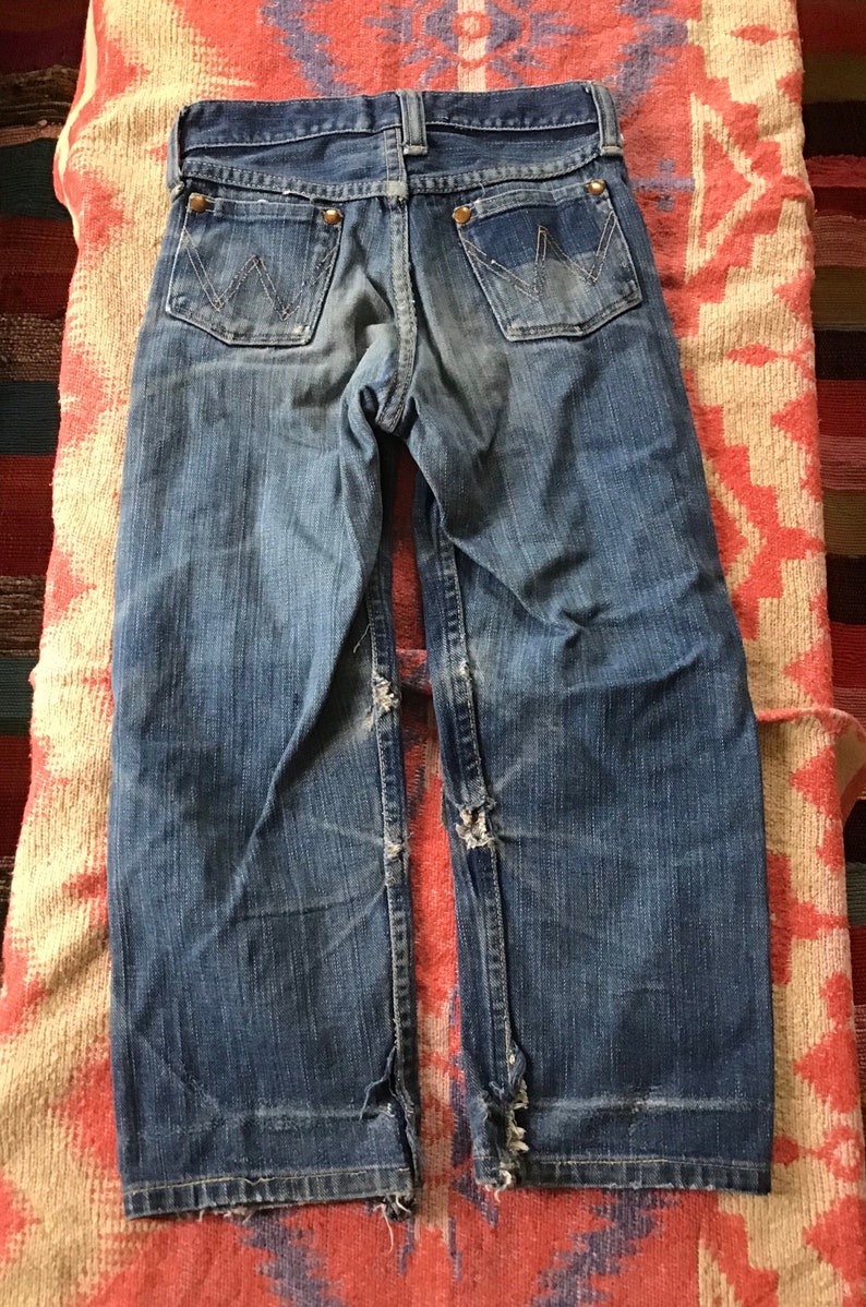 May include: Vintage blue denim jeans with the Wrangler logo on the back pockets. The jeans have a worn appearance with distressed details at the knees and hems. The jeans are laid flat on a patterned textile.