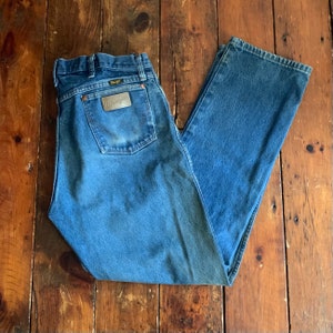 May include: Blue denim Wrangler jeans with a classic five-pocket design. The jeans feature a copper-colored leather patch on the back pocket with the Wrangler logo. The jeans are laid flat on a wooden surface.