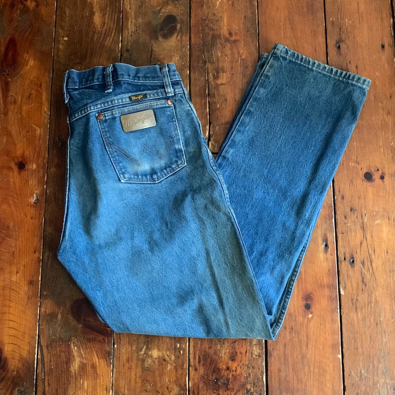 May include: Blue denim Wrangler jeans with a classic five-pocket design. The jeans feature a copper-colored leather patch on the back pocket with the Wrangler logo. The jeans are laid flat on a wooden surface.