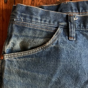 May include: Close-up of blue denim jeans, showcasing the pocket and waistband details. The jeans have a classic design with visible stitching and a copper-colored rivet. The fabric has a slightly faded appearance, suggesting wear.