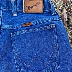 Pu&ograve; includere: Primo piano del retro di jeans blu. I jeans presentano una tasca posteriore con un design a 'W' cucito e un'etichetta in pelle marrone con il marchio 'Wrangler'. Le cuciture sono di colore beige a contrasto.