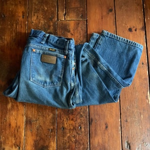 May include: Blue denim Wrangler jeans with a brown leather patch on the back pocket. The jeans are folded and laid on a wooden surface. The Wrangler patch is rectangular with the brand name in gold lettering.