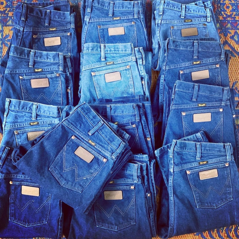 May include: A collection of blue denim jeans, folded and stacked. Each pair features a brown leather patch on the back pocket and the Wrangler logo. The jeans are in various shades of blue, from light to dark.