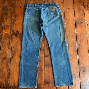 May include: Blue denim jeans with a classic five-pocket design. The jeans have a faded appearance and a brown leather patch on the back pocket. The jeans are laid flat on a wooden surface.