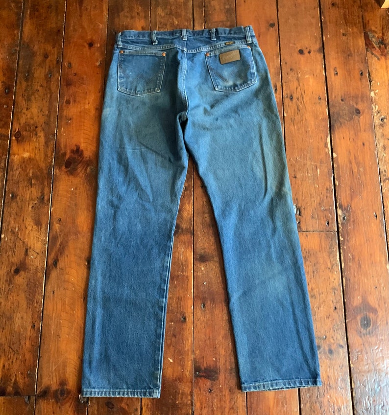 May include: Blue denim jeans with a classic five-pocket design. The jeans have a faded appearance and a brown leather patch on the back pocket. The jeans are laid flat on a wooden surface.