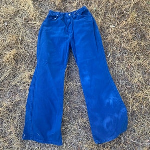 May include: A pair of vintage blue denim bell-bottom pants. The pants have a high waist and a wide flare from the knee down. The denim is a solid blue color with some lighter areas. The pants are laid out on a bed of dry grass.