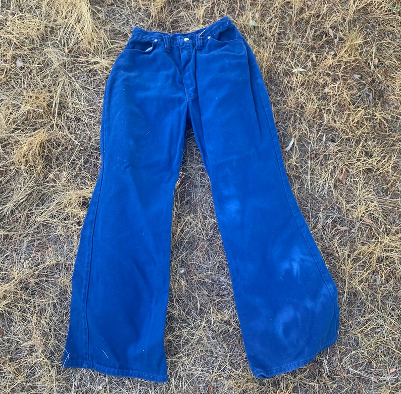 May include: A pair of vintage blue denim bell-bottom pants. The pants have a high waist and a wide flare from the knee down. The denim is a solid blue color with some lighter areas. The pants are laid out on a bed of dry grass.