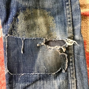 May include: Close-up of distressed blue denim jeans with a large patch sewn over a ripped area. The patch is a darker shade of blue, and the edges are frayed. The jeans show signs of wear and tear, with faded areas and a worn texture.