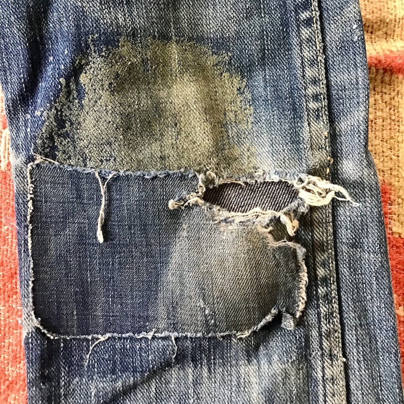 May include: Close-up of distressed blue denim jeans with a large patch sewn over a ripped area. The patch is a darker shade of blue, and the edges are frayed. The jeans show signs of wear and tear, with faded areas and a worn texture.