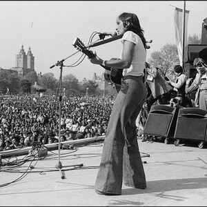 Pu&ograve; includere: Foto in bianco e nero di un musicista che si esibisce sul palco davanti a una folla numerosa. Il musicista suona una chitarra acustica e canta in un microfono. Il palco &egrave; all'aperto, con uno skyline sullo sfondo.