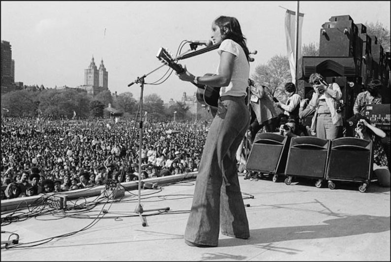 Pu&ograve; includere: Foto in bianco e nero di un musicista che si esibisce sul palco davanti a una folla numerosa. Il musicista suona una chitarra acustica e canta in un microfono. Il palco &egrave; all'aperto, con uno skyline sullo sfondo.