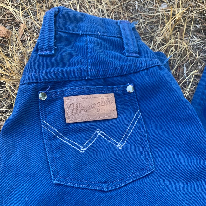 May include: Close-up of blue denim jeans with a back pocket featuring the Wrangler logo on a brown leather patch. The pocket has white stitching in a distinctive W shape. The jeans have silver rivets.