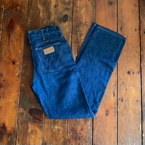 May include: Dark blue denim Wrangler jeans with a classic straight leg fit. The jeans feature a brown leather patch with the Wrangler logo on the back pocket. The jeans are folded on a wooden surface.