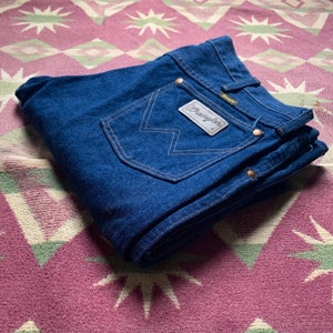 May include: Folded dark blue denim jeans with the Wrangler logo on the back pocket. The jeans have copper-colored rivets and a tan-colored stitching. The jeans are folded on a patterned, multi-colored textile.