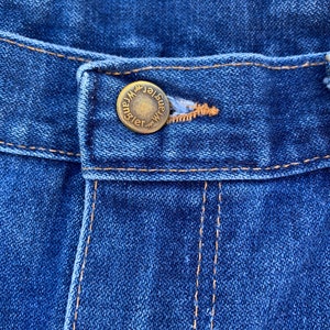 Pu&ograve; includere: Primo piano di jeans blu con un bottone in ottone. Il bottone &egrave; impresso con la scritta "Wrangler". I jeans hanno cuciture arancioni e un classico design a cinque tasche. Il tessuto ha un aspetto strutturato.