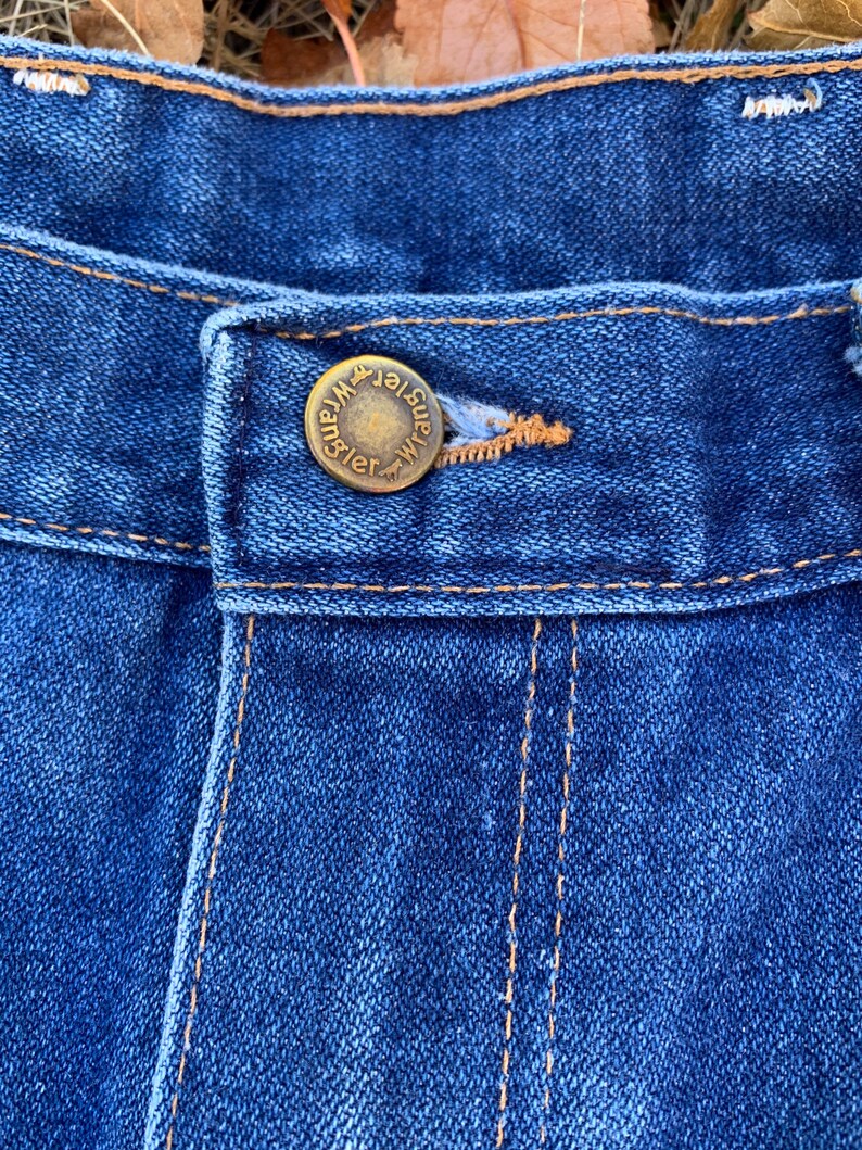 Pu&ograve; includere: Primo piano di jeans blu con un bottone in ottone. Il bottone &egrave; impresso con la scritta "Wrangler". I jeans hanno cuciture arancioni e un classico design a cinque tasche. Il tessuto ha un aspetto strutturato.