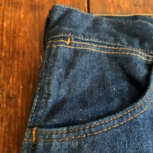 May include: Close-up of a section of dark blue denim jeans. The image highlights the pocket and stitching details. The stitching is a contrasting tan color, and the denim has a slightly worn appearance.