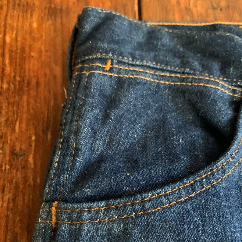 May include: Close-up of a section of dark blue denim jeans. The image highlights the pocket and stitching details. The stitching is a contrasting tan color, and the denim has a slightly worn appearance.