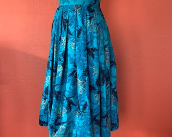 May include: A turquoise knee-length skirt with a pleated design, featuring a repeating pattern of dark blue and silver botanical motifs. The skirt is displayed on a mannequin, highlighting its full shape and high waist. The skirt's length is approximately 60 cm.